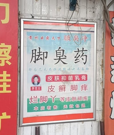 这是一张张贴在老旧墙面上的带金属边框的宣传海报，主打足部及皮肤问题治疗产品。海报顶部标注“贵州云霸天下 腋臭净”，核心位置用醒目大字突出“脚臭药”，搭配宣传语“用后几天不换洗鞋袜也不臭脚”吸引关注。海报中间印有创始人敖世云的正装头像，明确产品为“皮肤抑菌乳膏”，可针对皮癣、脚痒、烂脚丫等多种皮肤顽疾，底部以绿色区域标注“本店有售 无效包退”，做出售后承诺，整体风格平实接地气，是典型的市井小店线下宣传物料，用于招揽有相关皮肤困扰的顾客。
