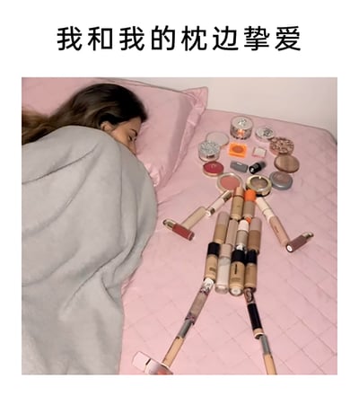 这是一张充满趣味的创意摄影图，画面中一位女性盖着灰色毯子，侧躺在粉色的床上熟睡。在她的枕边，创作者用粉底液、口红、眼影、腮红等各类化妆品精心摆放出了一个类似人形的造型，搭配图片上方的文字“我和我的枕边挚爱”，以幽默诙谐的方式调侃对于喜爱美妆的人群而言，化妆品就如同挚爱一般，是想要时刻放在身边的存在，精准戳中了美妆爱好者的心声。