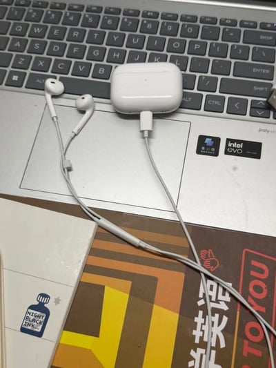 这是一张实拍的桌面场景图，画面上层是银灰色笔记本电脑的键盘区域和掌托，掌托上贴有intel evo认证标识和国产笔记本品牌标识，掌托位置放着白色的TWS无线蓝牙耳机充电盒，充电盒的充电口上插着一根Lightning接口的白色有线入耳式耳机的线，两个耳机头随意放在充电盒左侧的掌托上，耳机线向下延伸搭在笔记本下方的物品上；画面下层露出了笔记本下方放置的两本物品，左侧是印有“NIGHT BLACK INK”墨水瓶图案的米白色笔记本，右侧是封面带有黄棕色条纹和“学英语”字样的英语学习相关书籍。整体搭配违背常规电子产品使用逻辑，将原本无线使用的蓝牙耳机充电盒和有线耳机结合，形成了反差搞笑的效果。