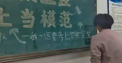 这是一张实拍的校园场景图，画面里一名背对着镜头的学生站在黑板前，黑板上写着「当模范」以及「我一定要考上研究生」的励志话语，同时用锤子图案和箭头设计了谐音梗，将「考上研究生」的表述和「上当」的暗示结合，形成反差感，搞笑又真实地体现出考研学生在备考时的复杂心态——一边喊着励志口号，一边又自嘲式地调侃自己“上当”投入考研大军。