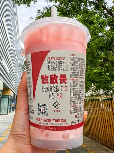 图片展示一杯粉色奶茶，但其外包装被制作为“敌敌畏”农药的仿真标签，内容包括“有效成分含量77.5%”、“剂型：乳油”等农药常见术语，仿佛手握农药桶但实际上却是饮品。包装和饮品之间表现出极大反差，极具恶搞色彩，通过字体设计和标签内容营造幽默、玩梗效果。

图中核心双关在于“敌敌畏”与“滴滴味”谐音，暗指奶茶有味道，以及借用农药标签反差吐槽。“敌敌畏”本身在网络上也常作为玩梗对象。
