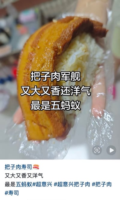 画面主体是一只戴着透明一次性塑料手套的手，托着一块体积很大、酱色红亮、肥瘦分层的把子肉；肉块下方垫着一团白米饭，整体被刻意摆成类似军舰寿司的造型。图片中央叠加白字黑边文字：“把子肉军舰／又大又香还洋气／最是五蚂蚁”。底部白色区域是社交媒体文案，写有“把子肉寿司🍣，又大又香又洋气，最是五蚂蚁”，并带有#超意兴、#超意兴把子肉、#把子肉、#寿司等话题标签。“五蚂蚁”谐音日语“うまい（umai）”，意思是“好吃、美味”；配合“军舰”这一日式寿司说法，形成又洋气又搞笑的美食梗。右下角可见播放和搜索图标，说明这是一段短视频或社交平台视频的截图。背景为虚化的室内环境，主要视觉焦点集中在把子肉和米饭上。