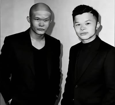 这是一张黑白单幅静态人像图，画面里有两名身着黑色西装、内搭黑色上衣的男性并排站立在纯色背景墙前，左侧男性为光头造型，双手插在裤兜中，右侧男性为短寸发型，两人都面向镜头，脸部特征几乎完全一致，是网友创作的玩梗内容，用来调侃网红许华升和广东雨神长相相似。