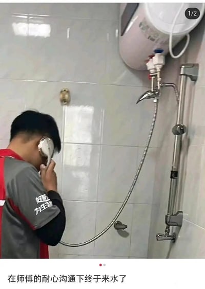 这是一张在卫生间拍摄的搞笑实拍图，画面中穿着维修工作服的师傅将刚安装好的淋浴喷头贴在耳边，做出打电话的姿势，配合图片底部的配文“在师傅的耐心沟通下终于来水了”，用拟人的幽默方式制造出“师傅在和水管沟通来水”的搞笑误会，实际是师傅在测试淋浴喷头是否正常出水，通过场景的错位感和文案的调侃，让普通的维修现场变得趣味十足。