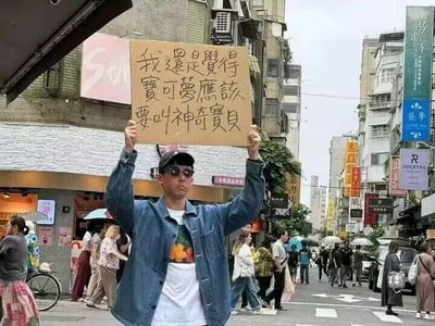 在台湾的一条城市街道上，一名戴着黑色墨镜和帽子、身穿牛仔外套的男子，双手举着一块手写的木牌站在路中央。木牌上写着“我还是觉得宝可梦应该要叫神奇宝贝”，直白地表达出他对宝可梦译名的个人偏好。周围是充满生活气息的街景，两侧分布着各类商铺，不少行人撑着伞在街道上路过，整体场景十分日常，没有双关、谐音梗或其他暗示内容。
