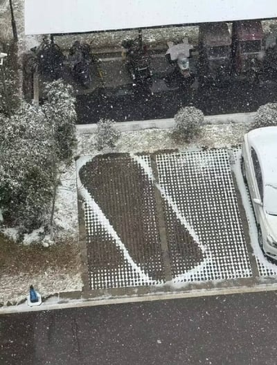 雪天的高空视角下，停车场呈现出一幅冬日景象。前景的停车区域被清理出独特的图案，雪被扫开后露出下方网格状的地面，形成类似几何设计的视觉效果。右侧停放着一辆被积雪覆盖的白色汽车，周围的灌木丛和地面都积着薄薄一层雪，雪花正纷纷扬扬地从空中飘落。背景中能看到一排停放在棚下的电动车或摩托车，整体场景静谧而富有生活气息，展现了普通人在雪天对停车空间的创意清理方式。