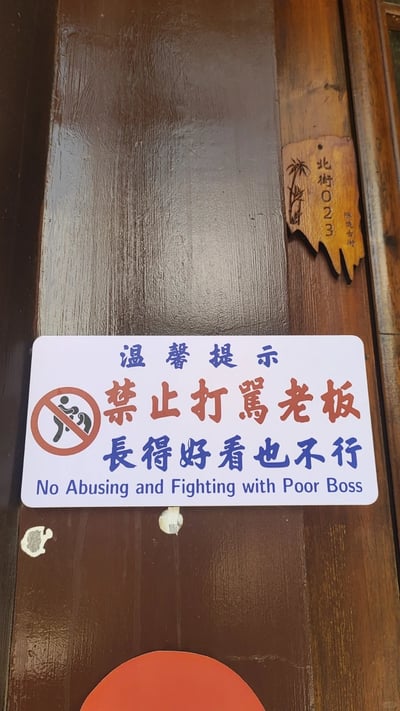 这是一张竖版原始实拍照片，画面主体是怀远古街北街023号店铺的深棕色木门区域。木门上贴有一块白底的趣味温馨提示牌：上方是蓝色字体的“温馨提示”，中间是醒目的红色字体“禁止打罵老板”，下方是蓝色字体的“長得好看也不行”，最底部配有英文翻译“No Abusing and Fighting with Poor Boss”，同时左侧还有一个代表禁止打骂的圆形禁令图标。提示牌右上方是一块边缘不规则的木质门牌号，上面刻有“北街023”字样，还有“怀远古街”的小字以及装饰性的植物图案。整个提示用幽默诙谐的语气，打破了常规提示的严肃感，增添了店铺的轻松氛围，十分有创意。
