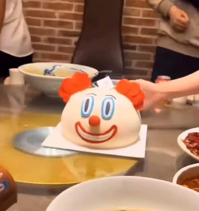 图片展示了一顿热闹的聚餐场景，餐桌上的火锅正冒着热气，而正中央摆放着一个造型夸张的小丑蛋糕成为视觉焦点。蛋糕以白色奶油为底色，顶部装饰着两撮鲜艳的红色卷发，蓝色的大眼睛和圆润的红色鼻子格外醒目，配上咧开的大嘴巴，表情十分魔性搞笑。一只手正伸向蛋糕，周围摆放着碗碟、调料和其他食物，背景是砖墙结构，整体氛围轻松欢乐，像是在庆祝某个特别的时刻。