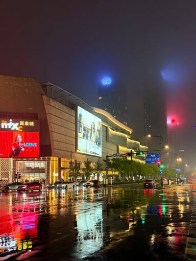 这是一张雨夜拍摄的城市街景实拍图，画面左侧是万象城商业综合体，外墙上设有LOEWE、DIOR的品牌广告大屏，建筑边缘有暖黄色灯带装饰。路面因降雨积水，清晰倒映出建筑灯光、广告牌色彩、交通信号灯等光源，倒影与实景呼应极具美感。远处的高层建筑被夜晚的雾气笼罩，顶部的标识灯光在雾气中产生朦胧的散射效果，整体画面的光影层次、雾气质感与3D渲染中使用体积雾+路径追踪技术产出的效果十分相似，观感梦幻高级。