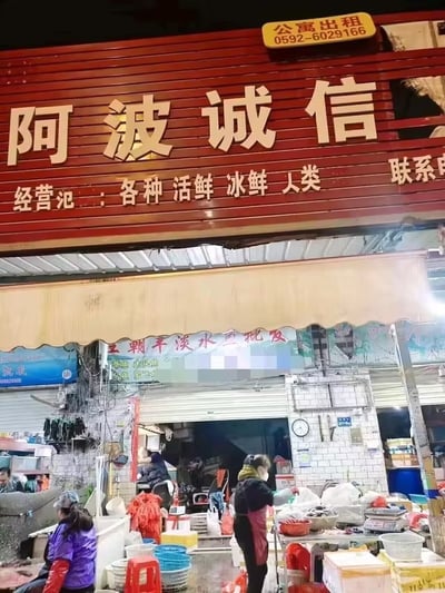 这是一家名为“阿波诚信”的水产档口，门头招牌写有“经营：各类活鲜 冰鲜 人类”，将“人类”列入经营范围，形成强烈的幽默和荒诞感。图中可见数名摊档工作人员正忙于处理海鲜、鱼类等，整体氛围是热闹的市场场景。主笑点和网络传播点在于“人类”二字的莫名出现，产生了黑色幽默和双关效果。

**双关/谐音梗说明：** 
- 明显是“人类”误写/误植，正常应该是“水产类”之类的词，但放在与海鲜并列处出现，造成令人发笑的错位感，广受网友恶搞和模仿。
