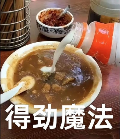 这是一张搞笑美食梗图，拍摄的是普通餐馆的桌面场景，碗里装着河南特色美食胡辣汤，有人正将橙白包装的娃哈哈营养快线乳饮料倒入胡辣汤中，桌面还摆放着盛有辣椒酱的小碗、金属筷子筒等常见餐饮用品，图片下方配有四个醒目的白色大字“得劲魔法”。内容是网友恶搞的猎奇黑暗料理吃法，将口味风格完全不兼容的两种食物混合玩梗，结合河南方言“得劲”的含义，增添了搞笑效果。