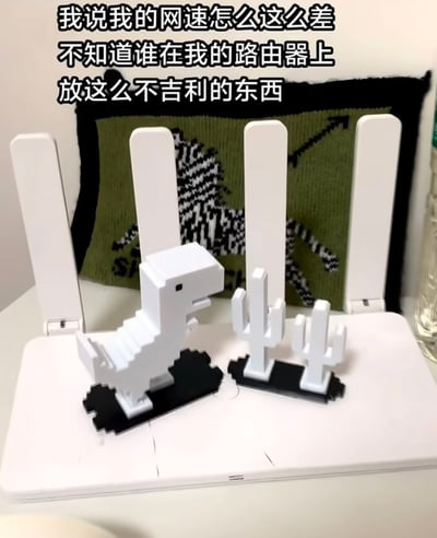 图片展示了一台白色多天线路由器，顶部放置着小型白色像素风格模型：一只恐龙和两株仙人掌。图片上方配有黑色文字：“我说我的网速怎么这么差 不知道谁在我的路由器上放这么不吉利的东西”。整体场景通过将路由器上的模型幽默地归咎为网速变差的“元凶”，营造出一种轻松搞笑的吐槽氛围，用生活化场景引发网友对网络问题的共鸣。