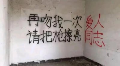 这是一张原始拍摄的静态图片，画面呈现了一个看起来被废弃的房间角落，墙面斑驳脱落，地面散落着杂物，整体环境显得破旧荒芜。墙面上有两处喷漆涂鸦：左侧墙面用黑色喷漆横向写着“再吻我一次”和“请把枪擦亮”两行文字，将浪漫的亲密请求与带有战斗感的指令结合；右侧墙角的墙面上用红色喷漆竖排写着“爱人同志”，这一表述兼具亲密关系与革命伙伴的双重意味，整体文字将柔情的浪漫与坚毅的力量感融合，营造出独特的革命浪漫主义氛围。