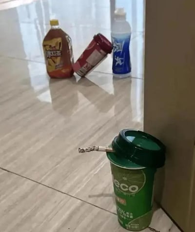这是一张利用巧妙拍摄角度制造视觉错位的趣味照片。前景中，一支点燃的香烟过滤嘴恰好对准绿色eco饮料杯的吸管口，从当前视角看，仿佛是这个饮料杯在“抽烟”；背景的光滑地面上，冰红茶、红色包装饮品、脉动三瓶饮料倾斜靠在一起，拟人化地像是在“围观”前景的饮料杯，整体画面通过视角错觉营造出充满脑洞的搞笑场景，将普通的物品赋予了拟人化的趣味行为。