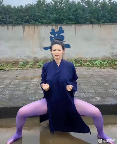 图中是一位身着深蓝色道袍、搭配紫色丝袜的女性，双腿大角度分开蹲在地面，双手握拳置于胸前。她身后的墙面上有一个蓝色的“莫”字，结合她分开双腿的视觉形态，恰好组成“墓”字，形成趣味视觉谐音梗。道袍的传统出尘感与紫色丝袜的现代感形成强烈反差，加上巧妙的文字视觉组合，让画面充满诙谐搞笑的氛围。