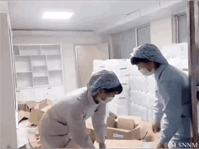 这是一段连贯的动态GIF，记录了工厂车间内发生的意外瞬间：两名身着工作服、佩戴防尘帽和口罩的工人在堆放着纸箱和大型容器的区域工作，其中一名工人抱着的大型容器突然脱手，他随即失去重心摔倒在地，容器内的液体瞬间倾泻而出，将地面和周围的纸箱浸湿打翻。另一名工人见状立刻上前，最后两人都在满是狼藉的现场，似乎在处理这场意外。整个过程充满了突发的慌乱感，直观展现了搬运操作中可能出现的安全事故。