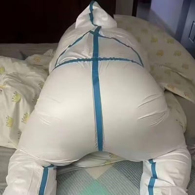 图片中展示了一个白色的充气或填充物体，其表面有蓝色的带子呈十字形交叉捆绑。该物体放置在一张床上，周围有几个带有黄色小动物图案的枕头。整个场景看起来像是在卧室环境中，光线较为柔和。物体的形状不规则，顶部有一个类似尖角的突起，整体呈现出一种被包裹或束缚的状态。
