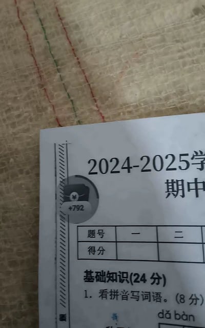 图片展示了2024-2025学年期中试卷的局部，可见卷首印有“2024-2025学年期中”字样，下方有题号（一、二）与得分的表格区域，以及“基础知识(24分)”模块，首题为“1. 看拼音写词语。(8分)”并标注了拼音“dǎ bàn”。试卷左侧边缘被添加了一个圆形图标，内含钱包图案及“+792”字样，形成幽默的反差感，疑似通过图片编辑软件将网络流行的“赚钱”元素与严肃的考试场景结合，制造调侃效果。