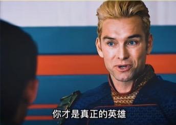 图片为电影剧照截图，画面中一名金发男性身着蓝色带有红色衣领的服装，正注视着镜头外的另一个人（仅可见对方后脑勺），背景为蓝橙相间的条纹幕布。图片底部有白色字幕：'你才是真正的英雄'，整体氛围诚恳且充满赞扬。