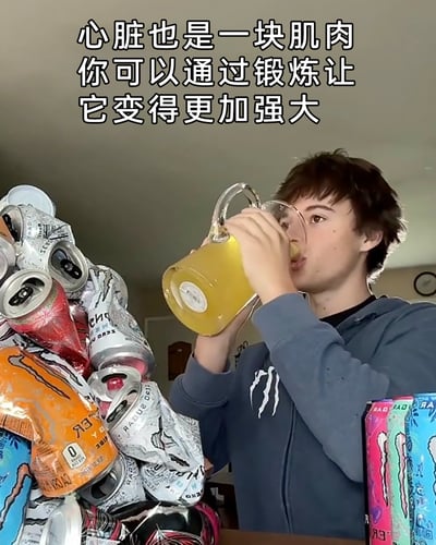 这是一张实拍的静态截图，图中一名穿着印有Monster（魔爪）标志连帽衫的年轻男性，正双手举着一个大玻璃壶，饮用里面的黄色功能饮料。他的身侧堆叠着大量被捏扁的Monster功能饮料罐，旁边还摆放着未开封的同款饮料罐。图的上方配有文字“心脏也是一块肌肉 你可以通过锻炼让它变得更加强大”，这里运用了反讽的恶搞梗——大量饮用功能饮料会给心脏造成极大负担，并非真正的“锻炼心脏”，用这种错误的、极端的行为来制造荒诞的笑点，属于网络上的整活内容。