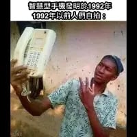 图中一位黑人男子将老式有线座机举至脸前，模仿现代人们用手机自拍的姿势，画面上方配有文字“智慧型手機發明於1992年 1992年以前人員自拍：”。这张图通过视觉错位和文字调侃，夸张地玩梗，将老式座机调侃为智能手机出现前的“自拍工具”，用幽默的方式对比现代自拍文化和早年的科技局限，制造出荒诞搞笑的效果。
