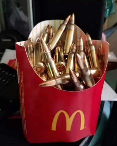 这是一张恶搞合成的静态图片，画面主体是印有麦当劳金色M标志的红色大号薯条包装盒，盒内没有装常规的薯条，而是塞满了大量黄铜色的步枪子弹，子弹弹头尖锐朝上摆放，形态与薯条原本的长条形态有相似性，制造出强烈的视觉反差和荒诞感，背景隐约可见键盘、桌面等物品，整体是将代表日常快餐的容器与具有杀伤力的武器结合的创意恶搞内容。
