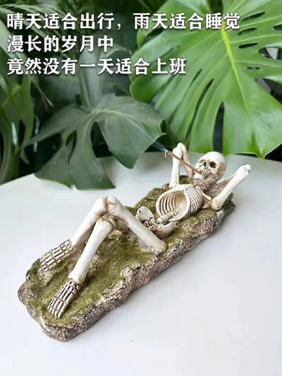 图片主体是一个平躺的骷髅摆件，它四肢舒展地躺在带有苔藓质感的底座上，嘴里叼着一根正在燃烧的香薰棒，背景是几片宽大的绿色植物叶子，营造出悠闲放松的氛围。图片上方配有文字：“晴天适合出行，雨天适合睡觉 漫长的岁月中 竟然没有一天适合上班”，以幽默的方式表达了对上班的抗拒心理，通过骷髅的“躺平”姿态与文字内容形成呼应，引发职场人士的共鸣。