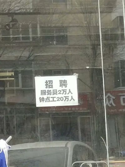 这是一张实拍的街边商铺玻璃门照片，玻璃门上贴着一张白色纸质招聘告示，告示用黑色字体写着“招聘 服务员2万人 钟点工20万人”，明显是店家书写失误：原本应该是标注薪资待遇（比如服务员月薪2万、钟点工时薪20元这类内容），错写成了招聘人数，出现了要一次性招聘两万服务员、二十万钟点工的离谱需求，完全不符合常理，十分滑稽；玻璃带有环境反光，可以看到对面建筑物和树木的倒影，玻璃下方能看到停放车辆的车顶，左下角只露出一点衣物边角，没有完整人物。