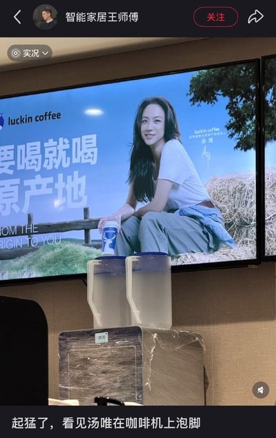 社交媒体用户“智能家居王师傅”发布的一张现场照片，画面中大型屏幕正在播放瑞幸咖啡代言人汤唯的广告，她身着白色上衣和灰色长裤，坐在干草堆上手持咖啡杯，背景是蓝天绿树，广告文案为“要喝就喝原产地”。屏幕正前方摆放着两个透明的方形水桶，因拍摄角度巧合，水桶与广告画面中的人物腿部形成错位，看起来像是汤唯正将双脚浸泡在水桶中，产生“泡脚”的趣味视觉错觉。图片底部配有文字“起猛了，看见汤唯在咖啡机上泡脚”，以幽默方式调侃这一巧合。