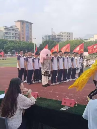 这是在学校操场举办的运动会活动现场，一排穿着统一蓝白校服的学生整齐地站在红色跑道上列队，其中一名学生戴着《孤独摇滚！》中后藤独（波奇酱）的粉色动漫头套，搭配浅色便装，和周围统一着装的同学形成强烈反差，显得格外有趣吸睛。前景处有一位长发女性（看起来像老师）坐在桌前，桌上摆放着写有“何敏”的红色名牌；画面右侧能看到舞狮表演的黄色狮尾部分。背景是绿色的操场草坪、围观的学生群体以及几栋教学楼，场地边缘的红旗上印有“高一”相关字样，整体氛围充满热闹活泼的校园活动气息。