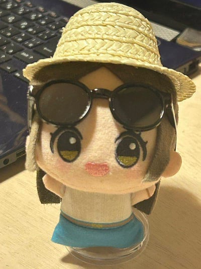 这是一张原始实拍图，主体是一个卡通风格的毛绒小玩偶。玩偶有着带有长睫毛的大尺寸黄绿色眼睛，粉色的微笑唇形，脸颊晕着淡淡的粉色腮红，自带软萌可爱的气质。用户为它额外搭配了浅米色的编织草帽和一副黑色大框墨镜，墨镜架在玩偶的眼睛上方，让原本软萌的玩偶多了一丝酷炫感。玩偶本身穿着米白色上衣和带有装饰边的蓝色短裙，被放置在一个透明圆形小底座上，背景是一台深色的笔记本电脑键盘，整体呈现出一种给玩偶打造夏日出行造型的趣味场景。