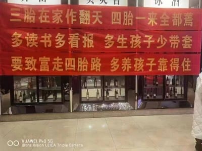这是一张实地拍摄的静态单图，画面主体是悬挂在一家酒类店铺门前的三条红色横幅，背景中可以看到摆放着各类酒瓶的玻璃展示柜，上方还有“冰酒”等店铺字样。横幅上用醒目的金色字体印着三段调侃式的鼓励生育标语，从上至下依次为“三胎在家作翻天 四胎一来全都蔫”“多读书多看报 多生孩子少带套”“要致富走四胎路 多养孩子靠得住”，这些标语采用口语化、夸张的表达，以戏谑的方式宣传多生育，其中部分标语内容较为直白。画面右下角露出了一位穿着白色外套的人的部分身体，左下角带有“HUAWEI P40 5G Ultra Vision LEICA Triple Camera”的拍摄设备水印。