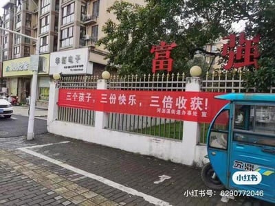 这是一张城市社区临街区域的实景拍摄照片，画面核心是白色金属围栏上悬挂的一条红色宣传横幅，横幅上印有白色文字：“三个孩子，三份快乐，三倍收获！”，落款为“河沥溪街道办事处 宣”，是街道办事处用于宣传三孩生育政策的正向引导标语。背景可见多层居民楼、临街商铺，包括“华拓电子”“川之味”的店铺招牌，围栏旁有树木，右下角停着一辆印有“苏鼎”字样的蓝色三轮车，地面是铺砖的人行道，整体呈现出日常平和的城市社区街景，没有双关、谐音梗或暗示类内容。
