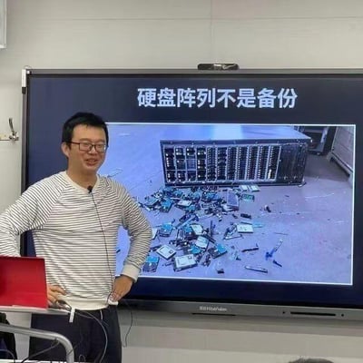 这是一张室内技术培训场景的实拍图，画面左侧是一位戴眼镜、穿条纹上衣的男士，他双手叉腰，面带笑容，正在进行一场数据存储相关的技术分享。他身后的大屏幕上显示着标题“硬盘阵列不是备份”，下方配了一张极具冲击力的图：一台完整的硬盘阵列设备旁，散落着大量损坏的硬盘和零件，用直观的视觉效果强调硬盘阵列无法替代备份的核心观点——一旦阵列损坏，数据将面临丢失风险，以此纠正技术认知误区。