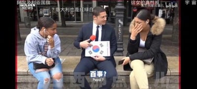 街头采访场景中，一名西装革履的记者手持韩国国旗坐在长椅中间，左右各有一位女性。左侧女性戴耳机、穿破洞牛仔裤，单手捂嘴笑；右侧女性穿黑色带毛领外套，双手捂脸显尴尬。画面中央有醒目文字“百事? Pepsi?”，幽默暗示将韩国国旗误认为百事标志。顶部叠加多条中文评论，如“阿尔及利亚国旗”“意大利也是红绿白”“这女的真性感”等，增添搞笑氛围。背景为城市街道，有建筑物和路灯。