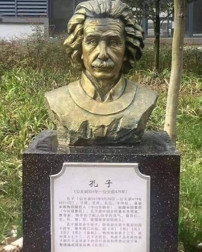 这张图呈现了一个充满违和感的拼接场景：画面上方是阿尔伯特·爱因斯坦的半身铜像，有着他标志性的蓬松卷发和严肃的面部神态，是这位科学名人的经典雕像造型；但下方的黑色基座铭牌上，却用大段文字详细介绍了中国古代思想家孔子的生平，包括孔子的生卒年、身份背景、思想成就、代表著作等完整信息，将两位分属不同时代、不同领域的名人错误搭配，形成强烈的反差感，属于恶搞性质的创作。
