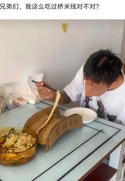 这是一张充满趣味的实拍图，图中男子为了玩“过桥米线”的字面梗，手工用硬纸板制作了一个写有“昆明大桥”的桥形道具，放置在玻璃餐桌上。男子将装着过桥米线的碗和空碗分别摆放在“桥”的两端，随后用筷子夹起米线，让米线从这个纸板‘桥’的上方‘穿过’，再送向嘴边，以此来字面化演绎‘过桥米线’的名称，制造出荒诞搞笑的效果。男子的脸部被打了马赛克，他正专注地完成这个‘过桥’吃米线的动作，桌面还能看到纸巾盒、小摆件等日常物品，整体场景是普通的室内餐桌环境。图片顶部配文‘兄弟们，我这么吃过桥米线对不对？’，进一步强化了玩梗的趣味性。