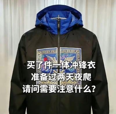 图中展示一件主体为黑色、连帽部分为蓝色的一体冲锋衣，衣服正面居中印有清代文官一品对应的仙鹤补子刺绣图案，图案外围还有传统回纹装饰，补子图案刚好被冲锋衣的中央拉链从中间分割开来。图上叠加了三行白色醒目文字，内容为“买了件一体冲锋衣 准备过两天夜爬 请问需要注意什么？”。这是一则搞笑玩梗内容，该补子是清代官服的标志性元素，也常出现在传统丧葬相关的服饰形象中，搭配夜爬的夜间户外场景，制造出穿这件衣服夜间出行容易被误认为是古代游魂或者古墓出土相关形象的荒诞搞笑反差，提问属于故意玩梗引导网友调侃回复。
