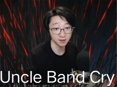 这是一张网友制作的网络梗图，主体是B站主播带篮子（陈义）的直播画面截图，人物戴着眼镜，面对麦克风，嘴巴微张，表情惊讶，似乎正在说话。画面背景被替换成黑红放射状的特效，底部配有英文文字“Uncle Band Cry”，是“带篮子哭”的谐音梗，用来调侃主播的相关情绪时刻，风格戏谑搞笑。
谐音梗说明：“Uncle Band”是“带篮子”的英文谐音，结合“Cry”组成玩梗内容，对应“带篮子哭”的调侃。