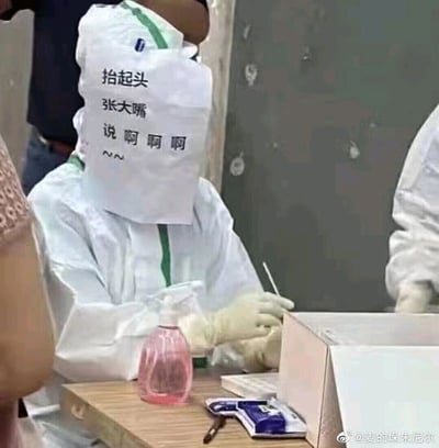 这是一张记录核酸采样场景的静态照片，画面中的核酸采样医护人员身着带有绿色线条的白色防护服，为了高效引导被采样人员配合操作，在防护面罩前张贴了一张白纸，上面写着“抬起头 张大嘴 说啊啊啊~~”的趣味提示语。医护人员戴着白色手套，手持采样拭子，桌上摆放着粉色的消毒喷壶，整个场景既体现了防疫工作的严谨性，又通过小巧思增添了趣味性，展现了防疫工作者的创意与贴心。