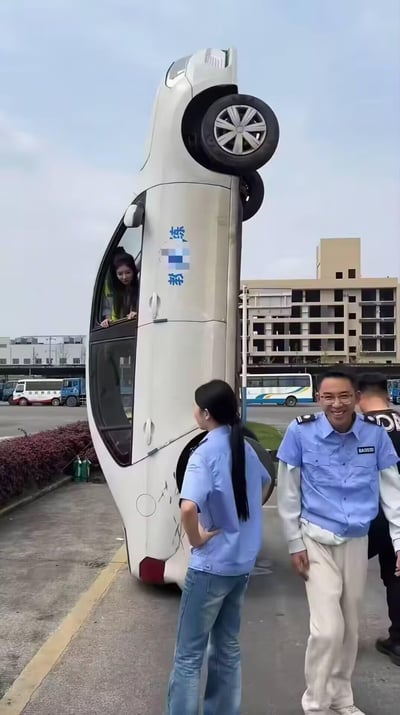 这是一张在驾校场地拍摄的视觉错位趣味照片：一辆印有“教练”字样的白色教练车，在特定拍摄角度下，看起来仿佛整个车身垂直立起，营造出学员把车开成直立状态的夸张错觉。一名女生从车窗探出头，地面上的几位驾校工作人员，一位笑着看向镜头，一位侧身看向“直立”的车，整体画面充满趣味，调侃了驾校学车的搞笑场景，实则是拍摄角度带来的视觉骗局。
