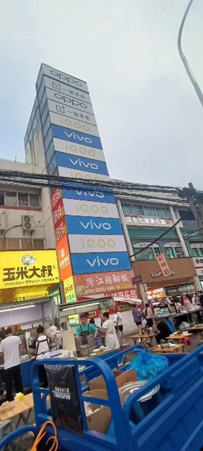 这是一张街头实拍照片，画面中心是一栋高耸的建筑，其外立面上密集重复排列着多个手机品牌Logo及文字，包括“OPPO”“二手手机”“iQOO”“vivo”等，这些标识以蓝、白、灰等颜色交替出现，形成强烈的视觉冲击。建筑下方是热闹的市场区域，有多个商铺招牌，如黄色的“玉米大叔”、红色的“隆江猪脚饭”等，街道上摆满了摊位，有商贩在忙碌，行人穿梭其中，呈现出典型的市井生活气息。天空为阴天，光线柔和，整体场景充满生活感与商业氛围。