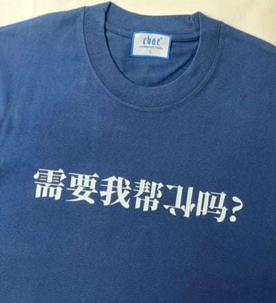 这是一件深蓝色圆领T恤的实拍图，T恤正面印有白色的玩梗文字「需要我帮ㄕㄐ吗？」，设计中将「忙」字拆分为「ㄕㄐ」（对应竖心旁的拼音首字母和「亡」的拼音首字母，同时也是字形拆分的视觉呈现），用拆字梗的方式把普通的问候语「需要我帮忙吗？」变得趣味十足，既保留了原本的热心问候含义，又增添了调侃、搞怪的氛围，适合作为潮流穿搭单品，传递轻松有趣的态度。