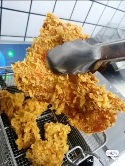 这张特写图展示了一块被金属餐夹夹起的炸鸡腿，其外皮是蓬松的鳞片状面衣，呈现出诱人的金黄色泽，看起来酥脆可口。下方的金属网架上还摆放着几块同款炸鸡，背景是厨房的操作区域，带有绿色指示灯的设备隐约可见，整体画面聚焦于炸鸡的诱人外观，充满了美食的治愈感。