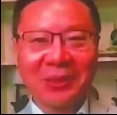 图片为一名戴眼镜男子的面部特写，面部呈现明显的泛红效果，背景模糊可见室内环境，可能是家具或书架。图像质量较低，有明显的像素感和色彩失真。