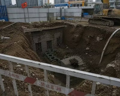 这是一张城市建筑工地的实拍照片，画面前景是带有“严禁抛物、禁止跨越”警示标识的施工防护栏，护栏后方是开挖出的大型土方坑，坑底露出了一处埋藏在地下的石砌老式建筑，建筑墙体上开有三扇加装了金属栅栏的窗户，建筑前方还排布有数个方形石制结构；土坑右侧停放着一台黄色挖掘机，土坑边缘设置有施工围挡，围挡后方可以看到城市高层建筑与临街商铺，整体呈现的是施工过程中意外发掘出地下未知老旧建筑遗存的现场场景。