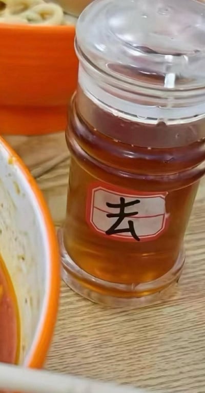 这是一张日常在外用餐场景的实拍照片，画面主体为摆放在浅木色桌面上的透明塑料调味瓶，瓶内装有棕红色的调味油，大概率为香油。瓶身贴有白底红边的标签，标签上仅手写了一个黑色的“去”字，看起来是原本要写的完整词汇只写了一半，显得莫名有喜感。画面左侧能看到部分吃剩的餐碗边缘，背景处还有橙色的餐盒，整体充满生活化的趣味感。