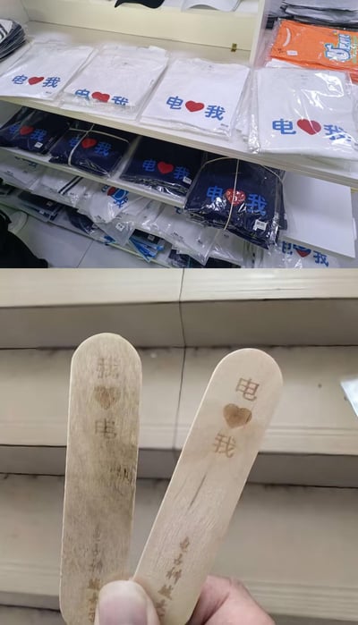这是一张上下拼接的实拍图，上图展示了货架上陈列的文创T恤，有白色和深蓝色两种款式，每件T恤都印着‘电❤️我’的字样，是‘电爱我’的谐音，带有电力相关的趣味属性；下图是两根木质冰棍棒，分别烫印着‘我❤️电’和‘电❤️我’的字样，和上方的T恤标语形成呼应，整体是电力主题的趣味文创周边，充满生活化的趣味感。