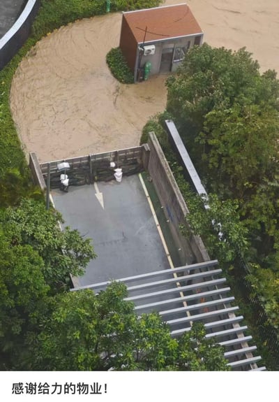 这是一张高空俯拍的实景照片，拍摄于暴雨引发城市内涝的场景下。画面下方是小区地下车库的入口坡道，入口处架设了金属挡水板，还堆叠了多层防汛沙袋进行加固，车库内部地面干燥，完全没有被水浸泡；画面上方的外部公共区域已经被浑浊的黄褐色洪水大面积淹没，一旁坐落着一间红棕色屋顶的小型保安岗亭，周边环绕着茂密的绿色植被。图片底部配有白色背景的黑色文字“感谢给力的物业！”，直观表达出对物业提前落实防汛措施、成功避免车库进水的肯定与赞许。