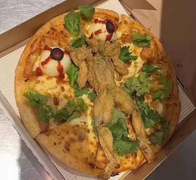 这是一款充满暗黑创意的猎奇披萨，放置在常规披萨盒中。披萨基底为烤至金黄的普通披萨，表面以一只完整的炸青蛙作为核心装饰，还搭配了用煮蛋、黑橄榄和番茄酱制作的仿真“眼球”造型，周围铺满香菜作为点缀，整体造型极具视觉冲击力，打破了常规披萨的外观设计，属于创意十足的暗黑料理，无谐音梗或双关含义。